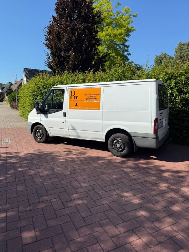 Weißer Transporter parkt auf einem gepflasterten Weg, umgeben von Bäumen und Sträuchern.